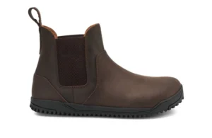A brown leather Chelsea Boot by Xero Shoes with a black rubber sole, elastic side panel, pull tab at the back, and minimal stitching, displayed from the side on a white background—ideal for those seeking a classic style and fit guide.